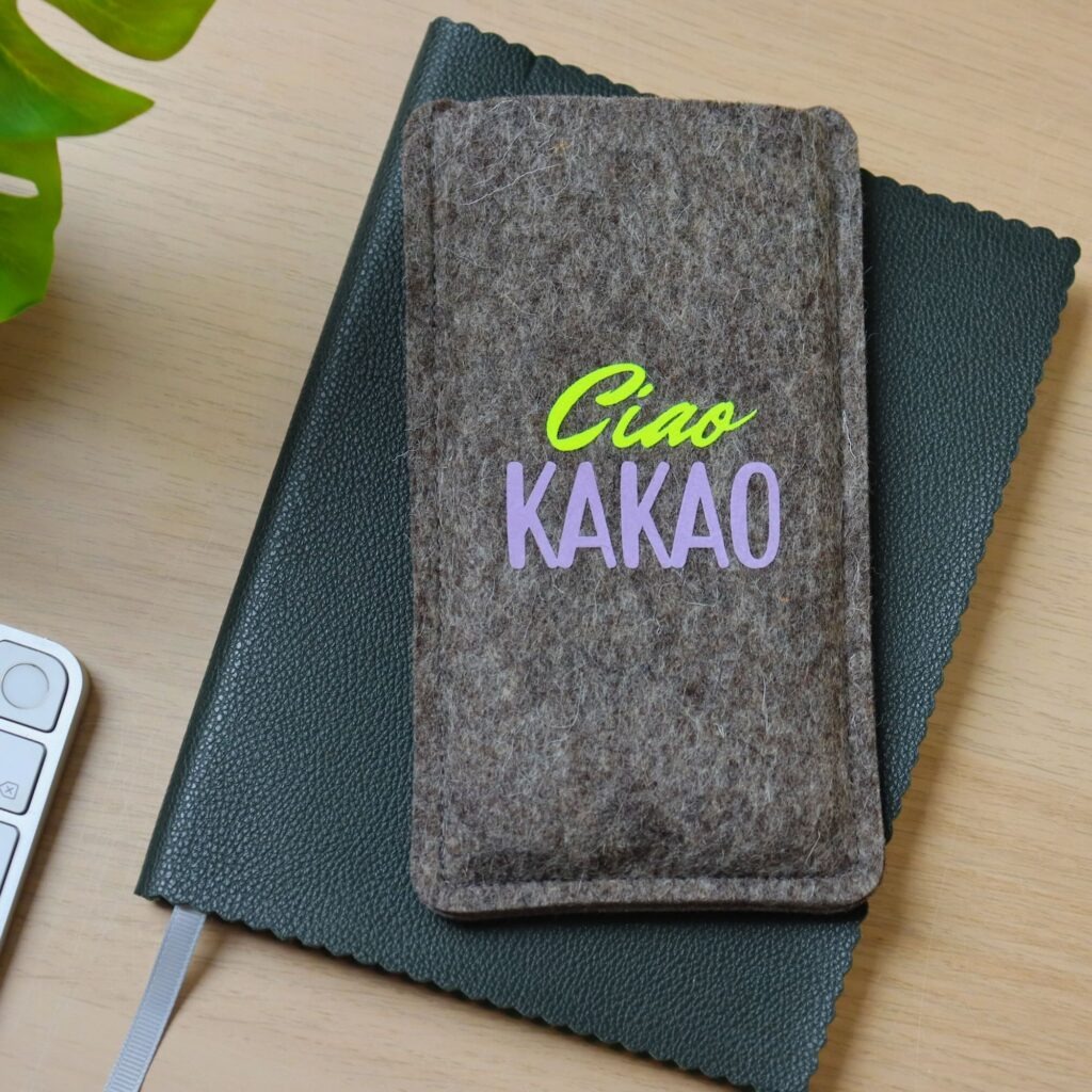 Handytasche aus grauem Filz mit Print "Ciao Kakao", auf einem Notizbuch liegend